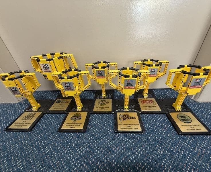 7 yellow trophies in the shape of a cup made from lego with black base and gold plaque with reason for award printed. Lego League logo on the cup
